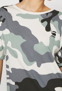 Lähikuva henkilöstä, joka käyttää lyhythihaista kamouflagekuvioista paitaa, jossa on vihreää, harmaata, mustaa ja valkoista väriä, sekä pieni "RAW"-logo rinnassa.