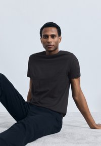 Tânăr cu păr creț scurt, purtând un tricou negru și pantaloni, așezat pe un covor gri deschis, lângă un perete alb simplu.