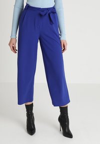 Pantalons larges bleus avec une taille à nouer, réalisés en tissu lisse. Associés à des bottes à talons hauts noires.