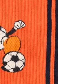 Κοντινό πλάνο πορτοκαλί πλεκτού υφάσματος με σχέδιο λευκής και μαύρης ποδοσφαιρικής μπάλας και ανυψωμένης γροθιάς, περιτριγυρισμένο από μαύρες και πορτοκαλί ρίγες.