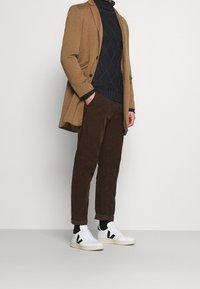 Uomo con cappotto color cammello, maglione blu scuro, pantaloni marroni e sneakers bianche con dettagli neri, in piedi su uno sfondo chiaro e uniforme.