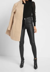 Cappotto color crema, top nero a maniche lunghe con texture, pantaloni in pelle nera aderenti e stivaletti neri con punta e cerniere.