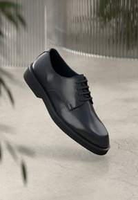 Μαύρο δερμάτινο παπούτσι με απαλή υφή, στρογγυλή μύτη, σχέδιο με κορδόνια και στερεά ελαστική σόλα, τοποθετημένο σε ουδέτερο φόντο.