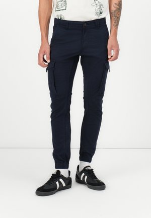 Marinblå cargobyxor med avsmalnande ben, elastiska mudd och sidofickor. Bärs med svarta sneakers med vita ränder och grå sulor.
