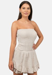 Strapless, lichtbeige jurk van gekreukeld materiaal. Heeft een aansluitende lijfje en een uitlopende, gerimpelde rok. Details omvatten een gestructureerde afwerking.