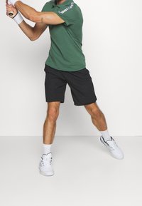 Man in green shirt and black shorts holds tennis racket, preparing to swing, wearing white sneakers and socks against plain background.