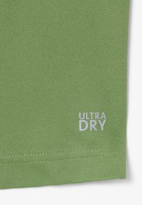 Tissu vert texturé avec ourlet doublement surpiqué et petit texte blanc « ULTRA DRY » imprimé près du bord.
