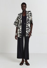Kimono-artiger Mantel in Schwarz- und Cremefarben mit Tierdruck, ausgestattet mit einem weit geschnittenen Ärmel-Design. Kombiniert mit einem schwarzen Top und weiten Hosen.