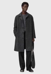 Grå uldfrakke med spidse kraver, knaplukning, sidelommer, båret over en struktureret grå sweater, kombineret med sorte bukser og en taske.