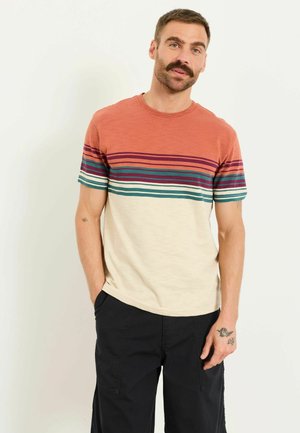 Mann mit Schnurrbart, der ein gestreiftes kurzärmeliges Hemd in Orange, Lila, Türkis und Creme sowie schwarze Hose trägt und mit einer Hand in der Tasche steht.
