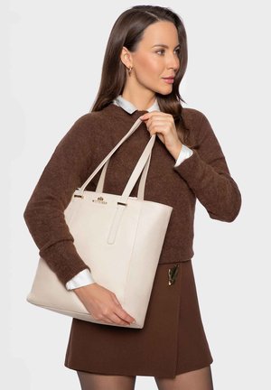 Kobieta w brązowym swetrze i spódnicy trzyma dużą beżową torbę Wittchen typu tote z podwójnymi ramiączkami, zwrócona w prawo.