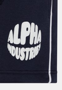 Tumši jūras zila auduma gabals ar baltu uzrakstu. Iekļauj apļveida logotipu ar vārdu "ALPHA" augšpusē un "INDUSTRIES" apakšpusē, rakstīts boldētā, noapaļotā fontā.
