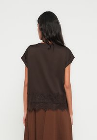 Καφέ μπλούζα με κοντά μανίκια, διακοσμημένη με δαντελένιο τελείωμα στο κάτω μέρος. Το μεταξένιο ύφασμα και η απαλή υφή δημιουργούν μια μαλακή εμφάνιση.