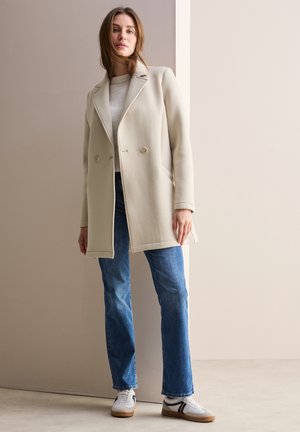 Beige Doppelrippenmantel mit Reverskragen, seitlichen Taschen, getragen über einem leichten Pullover und blauen geraden Jeans, kombiniert mit weißen Turnschuhen.