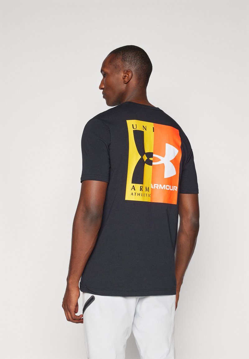 Crna majica kratkih rukava s višebojnom grafikom na leđima, koja sadrži žute, narančaste i crne elemente te bijele logotipove.