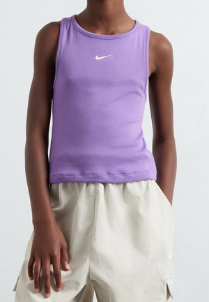 Person iført en ærmeløs lilla Nike-top og lyse beige shorts med hænder afslappet ved siden af kroppen på en ensfarvet baggrund.