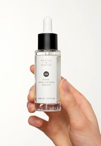 Bouteille compte-gouttes en verre transparent avec un capuchon noir, étiquetée "Pestle & Mortar Pure Hyaluronic Serum", contenant un sérum transparent. Main tenant la bouteille.