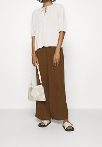 Světle růžová blůza s krátkým rukávem a zavazováním kolem krku; hnědé široké kalhoty; bílá tkaná taška s texturovaným vzorem; černé sandály s přezkou.