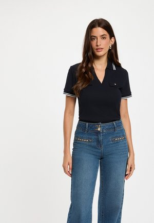 Frau mit langen braunen Haaren trägt ein dunkles Poloshirt mit weißem Besatz und hoch taillierte blaue Jeans mit dekorativen Fronttaschen.
