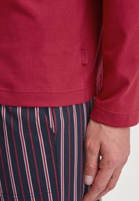 Rood overhemd met lange mouwen van katoen, met een subtiele textuur en een label aan de zijkant. Gecombineerd met marineblauwe en witte gestreepte broek.