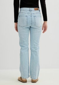 Jeans de tiro alto en un azul claro con corte acampanado y dos bolsillos traseros. Presenta una etiqueta de cuero marrón en la parte posterior de la cintura. Textura de denim suave.
