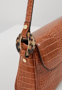 Bruine leren handtas met krokodillenpatroon, voorzien van een klep, gouden beslag en een cirkelvormige detail van schildpadmotief aan het hengsel.