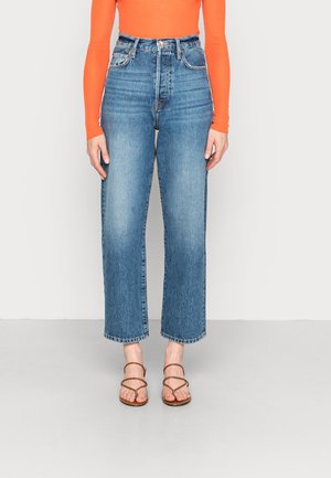 Kvinde iført højtaljede, lige ben blå jeans, orange langærmet top og brune sandaler med stropper, stående på en grå gulv.