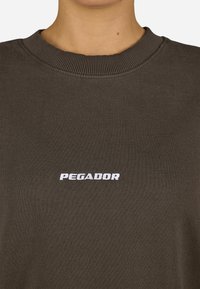 Brūns kokvilnas džemperis ar ribotu apkakli un baltu "PEGADOR" logo, kas centrēts uz krūšu. Gluda tekstūra un ikdienas dizains.