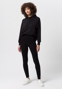 Černá mikina s kapucí, volného střihu a dlouhými rukávy, v kombinaci s černými legínami a bílými teniskami. Jednoduchý, monochromatický outfit.