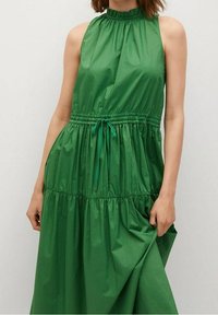 Zielona, bezrękawnikowa sukienka z wysokim dekoltem, marszczonym pasem i warstwową spódnicą. Wykonana z lekkiego materiału o gładkiej teksturze.