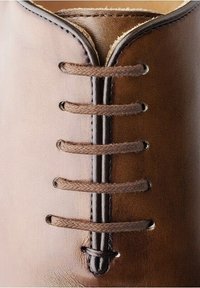 Dessus de chaussure en cuir marron avec une texture lisse, système de laçage fermé, et six œillets marron foncé avec des lacets tressés.