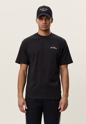 Jeune homme portant une casquette noire et un t-shirt noir avec des logos de texte blancs, debout devant un fond clair uni, faisant face.