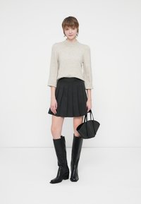 Suéter cinza claro, saia plissada preta, botas pretas até ao joelho, segurando uma pequena bolsa preta, em pé contra um fundo branco.