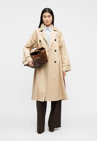 Γυναίκα που φοράει μπεζ παλτό τύπου trench πάνω από ανοιχτό μπλε πουκάμισο και σκούρο καφέ παντελόνι με φαρδιές φάσες, κρατώντας μεγάλη καφέ δερμάτινη τσάντα σε λευκό φόντο.