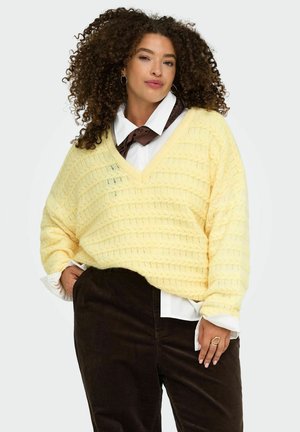 Femme aux cheveux bouclés portant un pull jaune en maille par-dessus une chemise blanche et un pantalon noir, posant les mains dans les poches devant un fond uni.