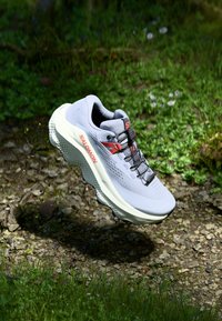 Chaussure de sport gris clair avec une tige en maille respirante, lacets noirs et accents rouges. Comprend une semelle blanche épaisse avec un grip texturé.