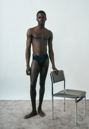 Slank mørkhudet mand stående barfodet i sorte Calvin Klein underbukser, hviler hånden på en grå stol med metalben mod en ensfarvet baggrund.