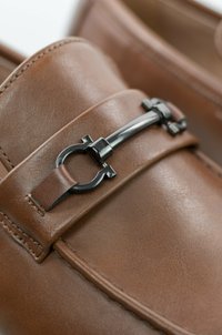 Mocasines de cuero marrón con un diseño elegante, que cuentan con un acento metálico y costuras detalladas a lo largo de los bordes. Textura suave en toda la superficie.