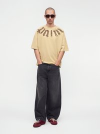 Homme portant un t-shirt graphique beige oversize, un jean noir large, des baskets bordeaux et des lunettes de soleil foncées, debout les mains dans les poches.