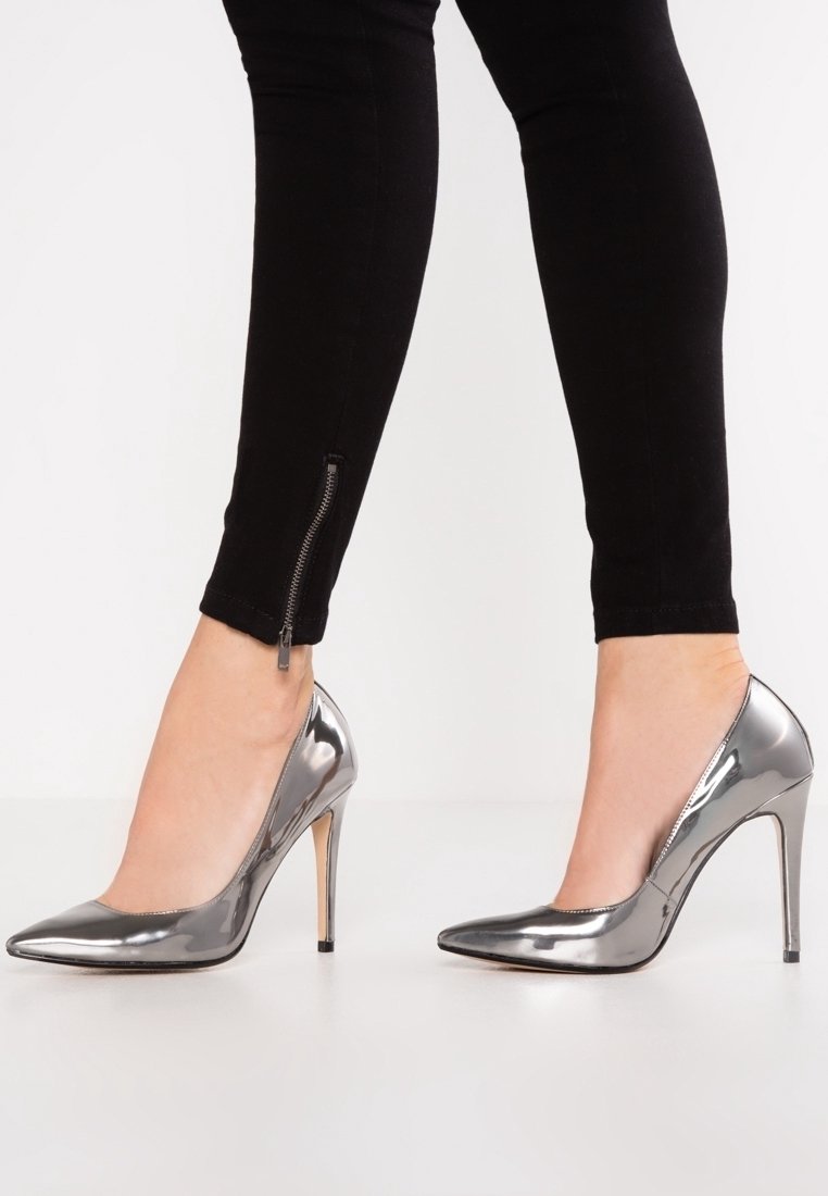 Jambes portant un pantalon slim noir avec des zips aux chevilles et des escarpins métalliques argentés à talons hauts et bouts pointus sur fond blanc.
