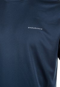 Sotilassi sininen urheilupaita, joka on valmistettu sileästä synteettisestä materiaalista, jossa on hienovarainen rakenne ja valkoinen "ENDURANCE" -logo rinnassa.