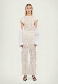 Combinaison en dentelle crème avec un motif géométrique, manches courtes à volants et détails sur les côtés. Associée à des manches longues blanches et des sandales noires.