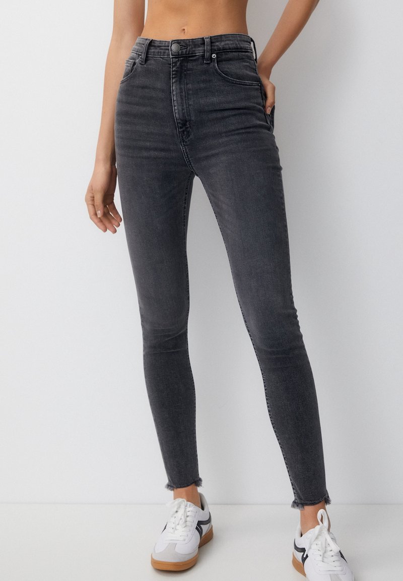 Jean slim taille haute gris foncé avec ourlets effilochés porté avec des baskets blanches à rayures noires sur une personne debout.