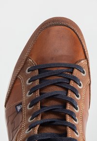 Brown leather shoe with navy laces, metal eyelets, and contrasting stitching. Features smooth and textured panels, with branding detail.