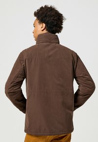 Giacca marrone leggera con colletto alto e maniche lunghe. Presenta una texture liscia e cuciture pulite lungo le cuciture.