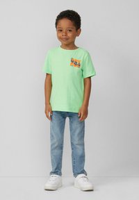 Groene katoenen T-shirt met korte mouwen en een "GET FUN" graphic in oranje en zwart, gecombineerd met lichtblauwe jeans en witte sneakers.