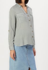 Donna che indossa una camicia a righe verticali verde e bianca con due tasche sul petto e una gonna di denim azzurro chiaro con uno spacco anteriore.