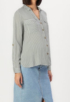 Donna che indossa una camicia a righe verticali verde e bianca con due tasche sul petto e una gonna di denim azzurro chiaro con uno spacco anteriore.