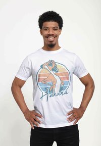 Hvid bomulds t-shirt med et grafisk motiv af en tegneseriefigur med teksten "Hakuna," sat mod et multikoloreret solnedgangsdesign.