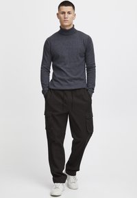 Mørkegrå turtleneck sweater parret med sorte cargo-bukser med sidelommer. Modellen står afslappet med hænderne i lommerne. Hvide sneakers på fødderne.